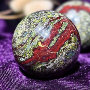May include: A polished stone sphere with a mix of colours. The sphere features a striking pattern of red, green, and white, with a marbled appearance. The stone is smooth and reflective, showcasing its natural beauty. A second sphere is slightly out of focus in the background.