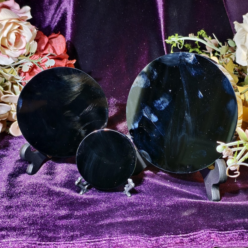 May include: Three black, circular scrying mirrors of varying sizes are displayed on small, black stands. The mirrors are set against a backdrop of a deep purple velvet cloth, with floral arrangements in the background.