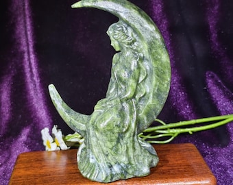 Può includere: Una scultura in pietra verde intagliata che raffigura una figura seduta su una luna crescente. La figura ha capelli lunghi e indossa un abito fluente. La pietra presenta motivi naturali maculati di colore verde scuro e nero.