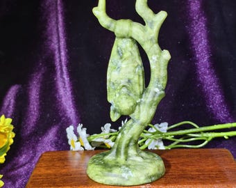 Può includere: Una scultura in pietra verde chiaro raffigurante un pipistrello appeso a testa in giù a un ramo d'albero. L'oggetto presenta una finitura liscia e lucida con venature minerali naturali, montato su una base circolare.