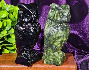 Può includere: Due statuette di gufo. Una è nera lucida, l'altra è verde con venature nere. Entrambe sono scolpite nella pietra e poggiano su una base di legno. I gufi sono uno accanto all'altro, mostrando la loro dettagliata maestria.