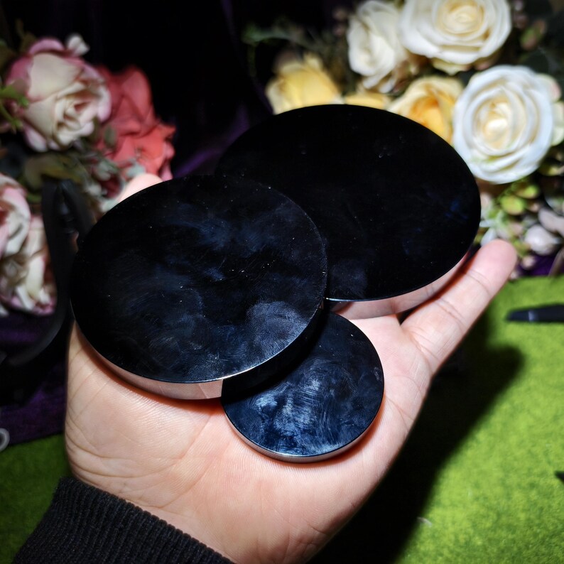 May include: Three black, circular objects with a reflective surface are held in a hand. The objects vary in size, with the largest at the top and the smallest at the bottom. The background features flowers.
