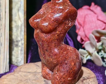 Può includere: Una scultura in pietra marrone-rossastra di un busto femminile. La scultura presenta seni definiti, una vita sottile e fianchi arrotondati. È esposta su una base di legno, con uno sfondo sfocato di fiori e altri oggetti.