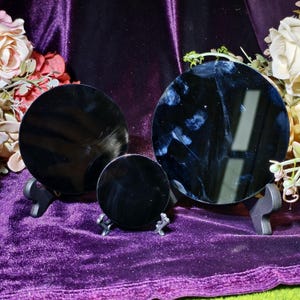May include: Three black, round mirrors of varying sizes are displayed on decorative stands. The mirrors are set against a backdrop of purple velvet fabric and accented with floral arrangements. The mirrors reflect light, creating a dark, reflective surface.