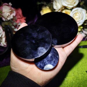 May include: Three dark purple, circular objects with a polished, reflective surface are held in a hand. The objects have a silver-colored rim. The background features a blurred arrangement of white and pink flowers.