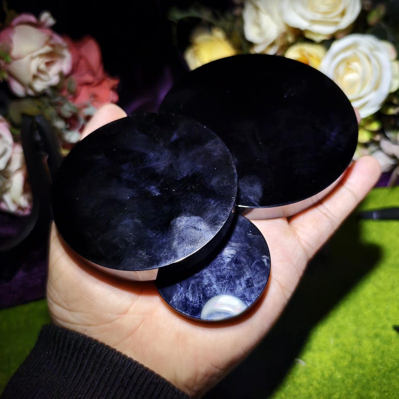 May include: Three dark purple, circular objects with a polished, reflective surface are held in a hand. The objects have a silver-colored rim. The background features a blurred arrangement of white and pink flowers.