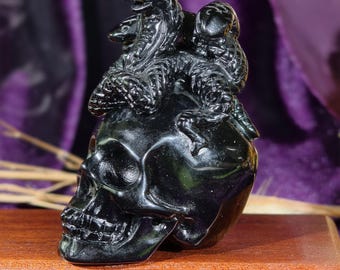 May include: A polished black obsidian carving of a human skull with three detailed snakes coiled around the top. The sculpture features a smooth, reflective surface with intricate scales on the snakes and defined anatomical features on the skull.