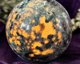 Può includere: Una sfera lucidata di labradorite con vivaci inclusioni arancioni, nere e blu. La gemma ha un aspetto maculato e una finitura lucida, che riflette la luce. Questa sfera decorativa è un oggetto di arredamento unico.