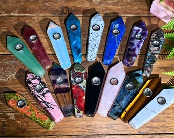 Può includere: Una collezione di sedici pipe in pietra levigata, ciascuna dotata di un inserto metallico cilindrico per il fornello. Le pipe presentano vari colori e motivi, tra cui verde, rosso, blu, nero, bianco, viola e marrone, disposte in modo sparso.