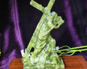 Può includere: Una scultura in pietra verde intagliata che raffigura una figura inginocchiata che trasporta una grande croce sulla spalla. La scultura presenta tonalità verdi chiare e scure screziate con una finitura lucida, su una base rettangolare.