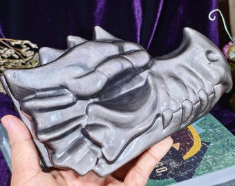 Può includere: Una scultura a forma di teschio di drago grigio metallizzato con denti affilati e dettagli. La scultura è tenuta in una mano, mostrando le sue dimensioni e il suo design intricato. Il teschio di drago è un oggetto decorativo.