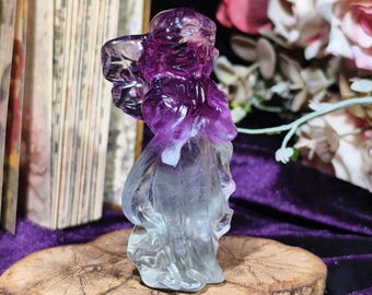 Può includere: Una statuetta scolpita di una persona con una veste fluente e ali, in un gradiente di viola e trasparente. La figura è esposta su una base di legno. Lo sfondo comprende un libro e fiori.