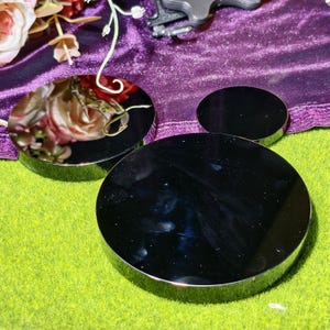 May include: Three black, round, reflective display stands with silver rims. The stands are arranged on a green surface, with a purple velvet cloth and floral arrangement in the background. The stands vary in size, with the largest in the foreground.