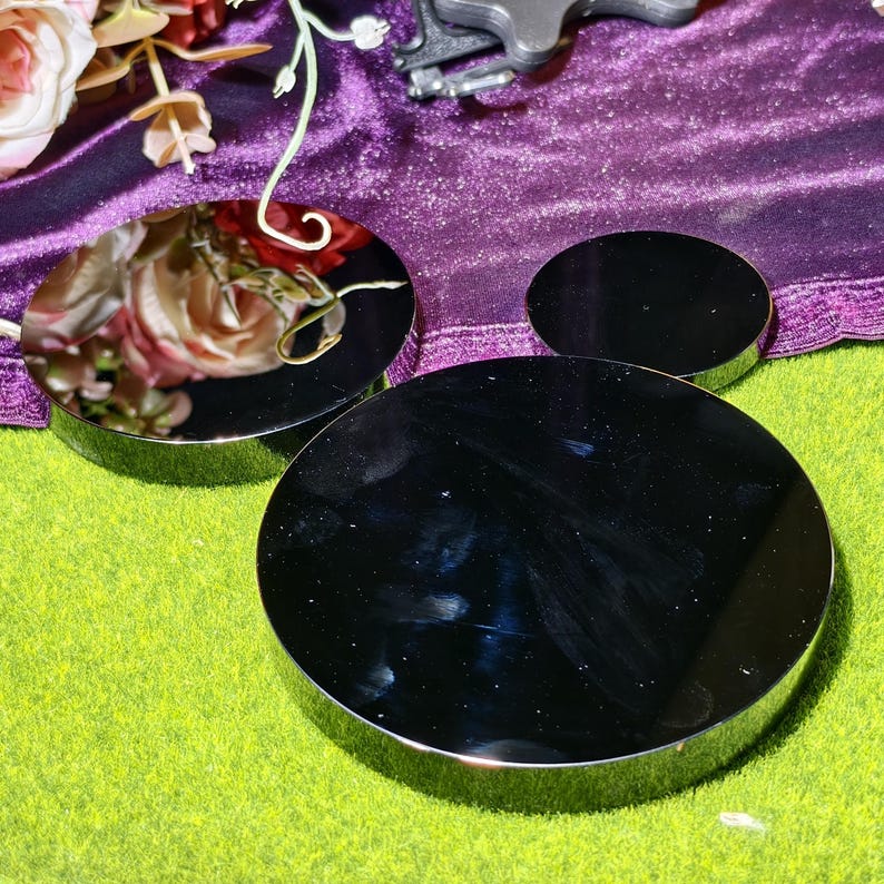 May include: Three black, round, reflective display stands with silver rims. The stands are arranged on a green surface, with a purple velvet cloth and floral arrangement in the background. The stands vary in size, with the largest in the foreground.