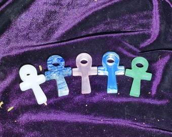 May include: Five ankh-shaped crystal pendants in white, blue, pink, light blue, and green. The pendants are displayed on a purple velvet surface. Each pendant has a loop at the top and a cross-like shape below.