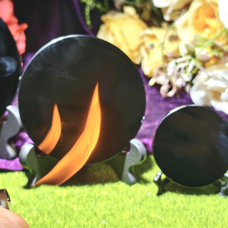 May include: Black, round, reflective objects with orange flame-like designs. The objects are displayed on decorative stands against a backdrop of flowers and a green surface. The objects appear to be decorative or artistic.