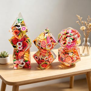 Op de afbeelding: Een verzameling heldere, geometrische dobbelstenen gevuld met kleurrijke fruitschijfjes en witte cijfers. De dobbelstenen zijn gerangschikt op een houten tafel, met een kleine vetplant en gedroogde bloemen op de achtergrond. De dobbelstenen zijn een uniek decoratief item.