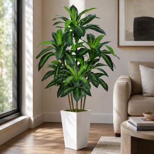 Puede incluir: Una planta artificial de Dracaena con hojas verdes y rayas amarillo-verdosas. Está en una maceta cuadrada blanca, sobre un suelo de madera. La planta añade un toque natural a la decoración de la habitación.