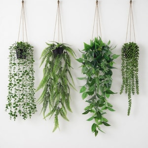 May include: Four hanging artificial plants with varying green foliage. Each plant is suspended by a brown rope from a white wall. The plants are in black pots, showcasing different leaf shapes and textures, creating a natural aesthetic.
