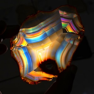 May include: A translucent, irregularly shaped gemstone with vibrant bands of color. The stone displays a spectrum of hues, including red, orange, yellow, blue, and green, creating a rainbow effect. The gemstone is held against a dark background.