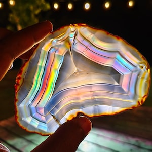 Peut inclure: Une tranche d'agate translucide présentant des bandes concentriques de couleurs blanches, grises et arc-en-ciel irisées. Le spécimen minéral possède un bord extérieur irrégulier et dentelé avec une fine bordure orange-rouge vive, éclairé par l'arrière.