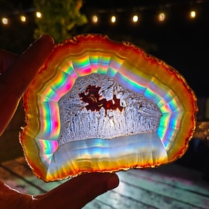 Peut inclure: Une tranche d'agate translucide présentant des bandes arc-en-ciel vibrantes et multicolores rayonnant à partir d'une cavité cristalline centrale texturée. Les bords sont définis par des teintes rouge et orange profondes, tandis que l'intérieur affiche un spectre de couleurs chatoyant et réfractant la lumière qui brille lorsqu'il est rétroéclairé.