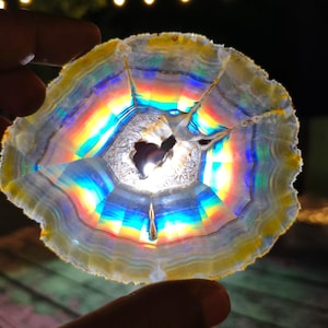 Peut inclure: Une tranche d'agate circulaire translucide présentant des bandes concentriques jaunes, blanches et bleues. La lumière traverse le centre, créant un effet arc-en-ciel prismatique vibrant à travers la structure cristalline et le cœur creux.