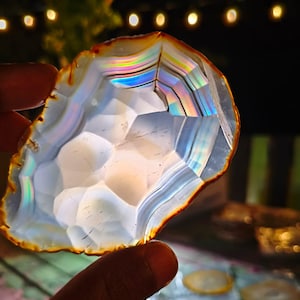 Peut inclure: Une tranche d'agate translucide présentant des bandes concentriques de couleurs blanches, grises et arc-en-ciel irisées. La pierre possède une texture de surface géométrique à facettes et un fin bord extérieur orange foncé laissant passer la lumière.