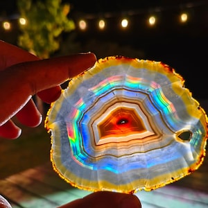 Peut inclure: Une tranche d'agate translucide présentant des bandes concentriques de couleur, dont un centre rouge profond entouré de couches de blanc, de jaune et de bleu. La pierre présente un effet prismatique arc-en-ciel vibrant avec un petit trou.