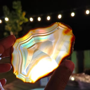 Peut inclure: Une tranche d'agate translucide présentant des bandes concentriques blanches, crème et ambrées. Le spécimen minéral est rétroéclairé, révélant un spectre arc-en-ciel irisé vibrant dans sa structure cristalline et un bord extérieur rouge orangé.