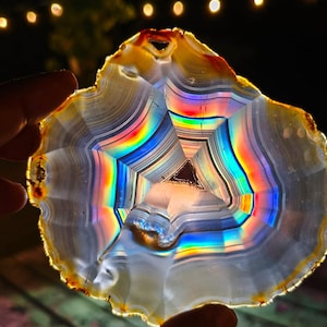 Peut inclure: Une tranche d'agate translucide présentant des bandes concentriques blanches, grises et ambrées, éclairée par l'arrière pour révéler un spectre arc-en-ciel vibrant de réfraction lumineuse à travers la structure cristalline et un vide triangulaire central.