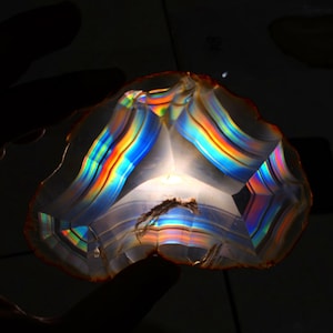 May include: A translucent, polished agate slice with vibrant rainbow iridescence. The gemstone displays bands of color, including red, orange, yellow, green, and blue, creating a stunning display of light refraction. The edges are rough and natural.