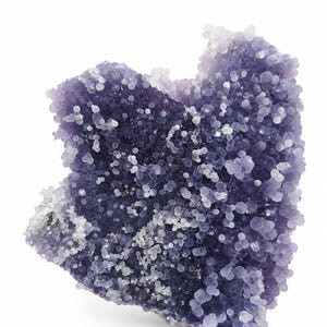 Puede incluir: Un espécimen mineral botrioide de color morado uva que presenta un grupo de pequeñas formaciones cristalinas redondeadas y translúcidas. La superficie está densamente cubierta de diminutas esferas que recuerdan a burbujas, con tonos desde violeta intenso hasta bordes blancos escarchados.