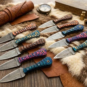 Può includere: Un assortimento di coltelli artigianali con manici decorativi in vari colori, tra cui blu, rosa e marrone. Le lame presentano un motivo unico. I coltelli sono disposti su una superficie strutturata con dettagli in pelle.