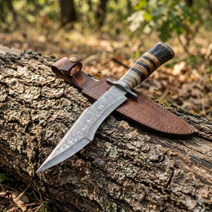 Könnte beinhalten: Ein Jagdmesser aus Damaststahl mit gemusterter Klinge liegt auf einem Baumstamm, neben einer braunen Lederscheide. Der Griff hat ein geschichtetes Design aus dunklen und hellen Tönen. Das Messer ist für den Außeneinsatz konzipiert.
