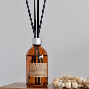 May include: An amber glass diffuser bottle with black reeds. The bottle has a silver cap and a label that reads "Velas - Verona." A wooden bead garland rests on a wooden surface next to the diffuser. The diffuser is a home fragrance product.