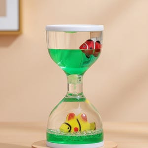 May include: Liquid motion toy in an hourglass shape, featuring two toy fish. One fish is red and white, the other yellow. The liquid is green, and the base is made of wood. The toy is designed to provide a calming visual effect.