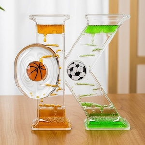 May include: A clear liquid motion timer in the shape of the letters O and K. The O features a basketball, and the K has a football graphic. The liquid is orange and green, creating a visual effect as it flows through the timer.