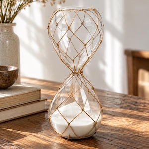 May include: A clear glass hourglass featuring a decorative tan twine netting wrapped around the exterior. The bottom bulb is filled with fine white sand, while the top bulb is empty. The hourglass has a rounded shape and a narrow centre neck.
