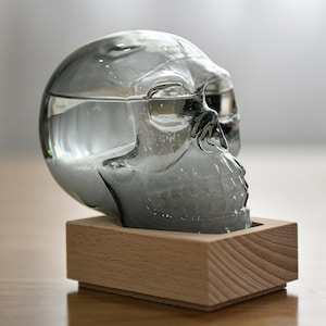May include: A clear glass skull-shaped decanter filled with water, set on a small, light-coloured wooden base. The skull has detailed facial features and is partially filled with liquid. The design is modern and minimalist.