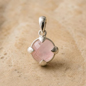 May include: A silver pendant featuring a round, faceted, pale pink gemstone set in a four-prong metal mounting. The setting has a polished, reflective finish with a small, integrated loop at the top for attachment to a chain or cord.