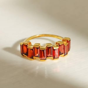 May include: A gold ring featuring a row of seven rectangular, baguette-cut deep red garnet gemstones set horizontally. The band is thin and polished gold, with each stone held in place by individual gold prongs, creating a continuous line.