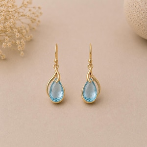 May include: A pair of gold drop earrings featuring faceted, pear-shaped light blue gemstones. Each earring is suspended from a curved gold wire hook, with the stone cradled by a delicate, sweeping gold metal frame that mimics a teardrop shape.