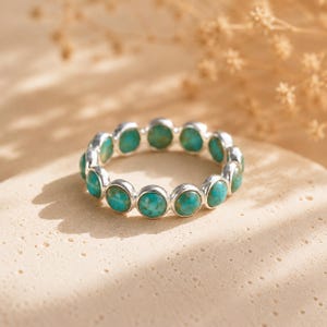 May include: A silver ring featuring a series of turquoise stones. The ring's band is a polished silver, and each turquoise stone is set in a bezel setting. The ring is displayed on a neutral-toned surface, with soft lighting.