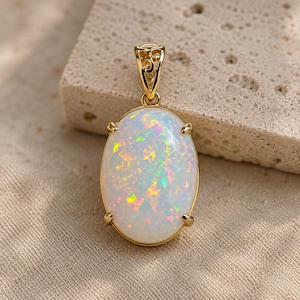 Könnte beinhalten: Ein ovaler Anhänger mit einem weißen Opal, der schillernde Blitze in Rosa, Grün und Gelb aufweist. Der Edelstein ist in eine polierte, goldfarbene Metallfassung mit vier Krappen und einer dekorativen Filigranöse an der Oberseite gefasst.