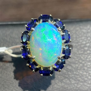 May include: A silver ring featuring a large, oval-shaped cabochon opal with vibrant green and orange play-of-color, surrounded by a halo of twelve deep blue, oval-cut faceted gemstones set in a prong setting on a metallic band.
