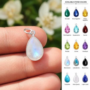 Könnte beinhalten: Ein tropfenförmiger Mondstein-Anhänger mit einer silbernen Öse. Das Bild enthält eine Tabelle mit dem Titel AVAILABLE STONE COLORS, die fünfzehn Edelsteinoptionen zeigt: Emerald, Blue Sapphire, Yellow Sapphire, Pink Sapphire, Aquamarine, Garnet, Peridot, Citrine, Amethyst, Moonstone, Labradorite, Black Onyx, Turquoise, Rose Quartz und Lapis Lazuli.