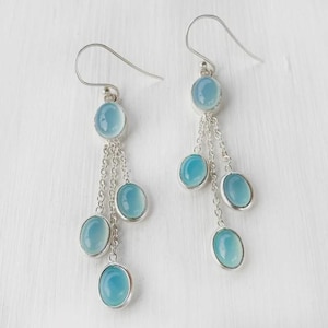 Puede incluir: Un par de pendientes colgantes con ganchos y cadenas de metal plateado. Cada pendiente presenta varias piedras de calcedonia azul claro, lisas y ovaladas, engastadas en metal, creando un diseño en cascada que cuelga verticalmente.