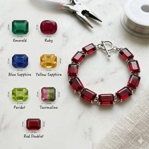 Op de afbeelding: Een armband met rechthoekige rode edelstenen en een zilveren sluiting. De afbeelding toont ook verschillende edelstenen: smaragd, robijn, blauwe saffier, gele saffier, peridot, toermalijn en rode doublet. Afmetingen in mm.