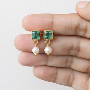 May include: A pair of gold-toned drop earrings featuring a square-cut, faceted green gemstone at the top. A small, round white pearl is suspended from each square gemstone by a thin gold-toned circular link, creating a delicate dangling design.